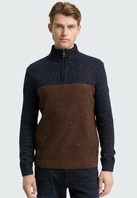 Junger Mann, der einen navyblauen und braun gesprenkelten Strickpullover mit kurzem Reißverschluss und gerippten Bündchen trägt, vor einem hellgrauen Hintergrund steht.