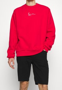 Felpa rossa con logo ricamato bianco sul petto, scollatura rotonda, maniche lunghe e vestibilità morbida. Indossata con pantaloni corti neri.