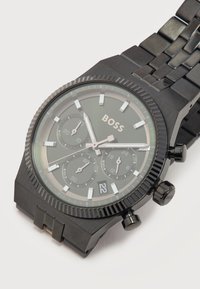 Μαύρο μεταλλικό ρολόι χειρός με καντράν χρονογράφου, ένδειξη ημερομηνίας που δείχνει 22 και λογότυπο "BOSS" στο σκούρο καντράν με μπρασελέ από μεταλλικούς κρίκους.