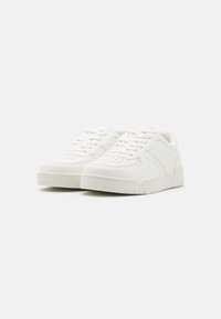 Weiße Sneaker mit einem strukturierten Leder-Obermaterial, perforierter Zehenbox, flachen Schnürsenkeln und einer dicken Gummisohle. Subtile Seitendetails werten das Design auf.