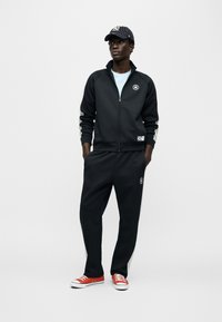 Μαύρο τζάκετ με φερμουάρ και λογότυπο, γκρι ρίγες στο πλάι, ταιριαστό παντελόνι και κόκκινα αθλητικά παπούτσια. Υλικό με υφή και άνετη εφαρμογή. Μαύρο και λευκό καπέλο.