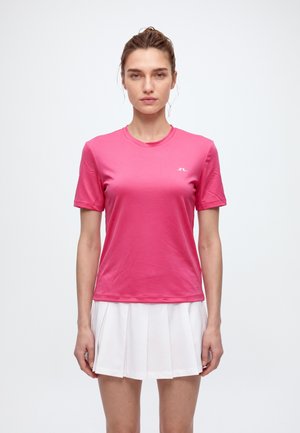 Une femme porte un tee-shirt de sport rose vif à manches courtes et une jupe plissée blanche sur un fond clair et uni.