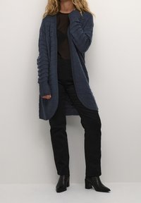 Cardigan lavorato a maglia blu navy con maniche a coste e texture, design aperto davanti, abbinato a pantaloni neri aderenti e stivaletti.