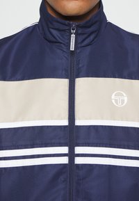 Navy zip-up jacket with beige and white horizontal stripes, featuring a logo on the chest and a high collar design. Textured fabric.