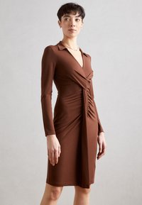 Robe longue marron à manches longues avec un col, présentant un design tordu à la taille et une silhouette ajustée, fabriquée en tissu extensible.