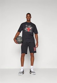 Čierne grafické tričko s červeným dizajnom býka a doplnkami s palmami, v kombinácii s čiernymi šortkami s nápisom "BULL" v červenej farbe. Model drží basketbalovú loptu.