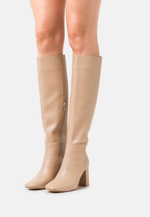Jambes portant des bottes en cuir beige hauteur genou avec talons blocs et fermetures éclair latérales sur un fond blanc uni.