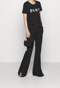 Svart DKNY t-shirt med en vit logo, kombinerat med svarta utsvängda byxor. En liten svart väska med kedjesnodd hålls i höger hand.