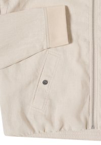 Manche et devant de veste beige avec poignet côtelé, poche à rabat inclinée avec bouton-pression métallique, et fermeture éclair visible sur le côté droit.