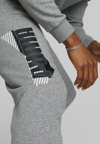 Des sweatpants gris présentent un logo noir au design angulaire et des bandes blanches, fabriqués à partir d'un tissu doux et texturé.