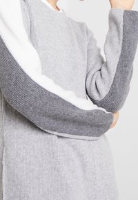 Suéter de punto gris con un cuerpo texturizado y mangas acanaladas en tonos gris claro y oscuro con detalles en blanco.