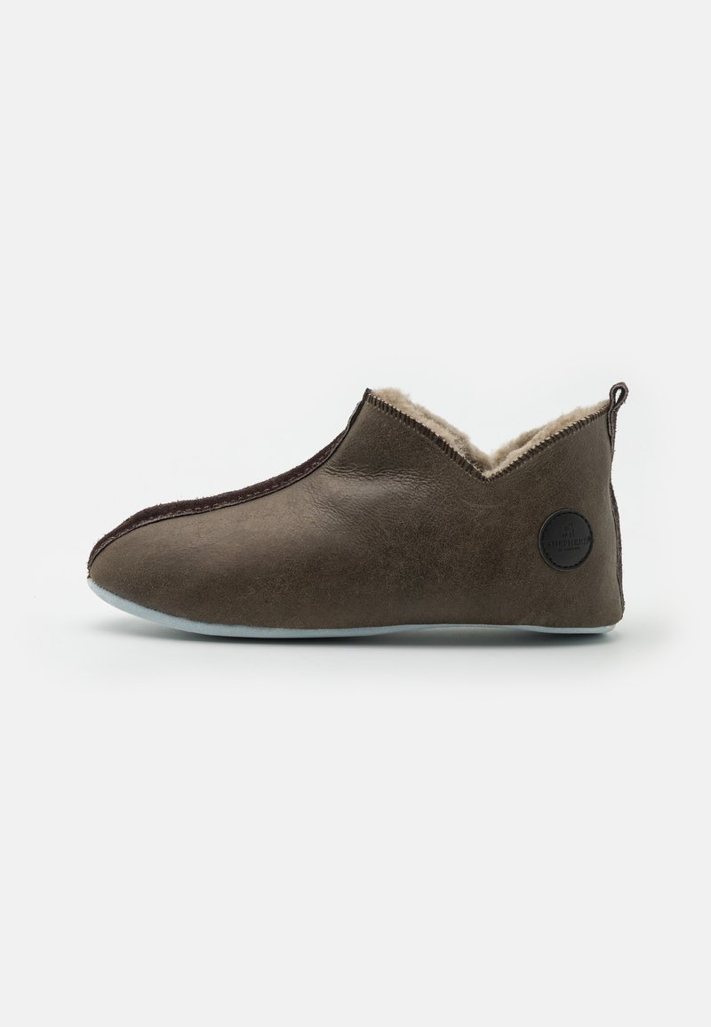 Brune læderslipper med blød, cremet foring, elastisk side og et sort cirkulært emblem på hælen. Flad, fleksibel sål.