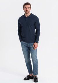 Man med händerna i fickorna, klädd i en marinblå långärmad polo, blå jeans och svarta läderskor, som står mot vit bakgrund.