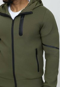 Sweat à zip olive avec une coupe ajustée. Présente des zips noirs, une capuche et des poches latérales. Texture lisse avec des accents minimalistes.