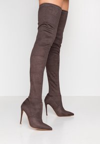 Stivaletti alti fino alla coscia, in suede marrone scuro con punta affilata, indossati da una persona in piedi su uno sfondo chiaro.