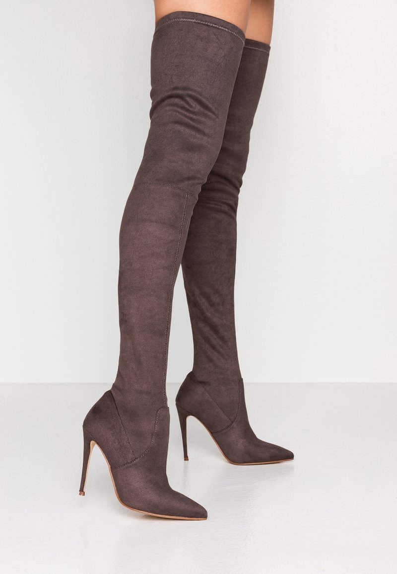 Stivaletti alti fino alla coscia, in suede marrone scuro con punta affilata, indossati da una persona in piedi su uno sfondo chiaro.