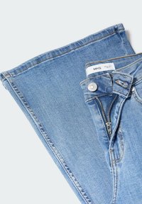 Jean en denim bleu clair avec fermeture éclair partiellement ouverte, bouton argenté et étiquette blanche visible à l'intérieur de la ceinture sur un fond uni.