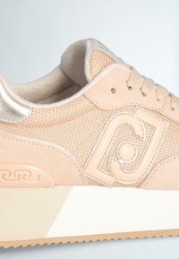 Sneakers beige con materiali in rete e camoscio. Presentano un colletto imbottito, accenti metallici, dettagli con logo e una suola in gomma bianca con texture.