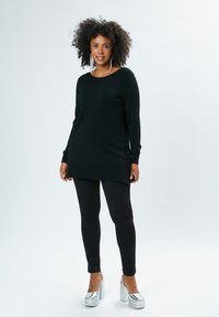 Tunika nera a maniche lunghe in maglia, con bottoni laterali, abbinata a leggings neri e tacchi a piattaforma argento. Tessuto liscio, vestibilità comoda.