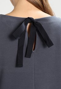 Harmaa paita pyöreällä kaula-aukolla, jossa on takana avaus, joka on kiinnitetty mustilla nauhoilla. Sileä pinta, rento muotoilu.