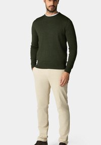 Olivově zelený svetr, kulatý výstřih, dlouhé rukávy, pletená textura, zkombinovaný s béžovými chinos kalhotami a tmavými botami, stojící na neutrálním pozadí.