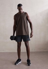Mand i atletisk tøj, der står indendørs, holder en dumbbell i højre hånd og kigger ned på vægten mod en ensfarvet baggrund.