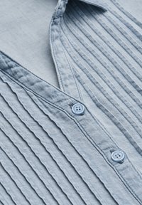  Lichtblauwe shirt met verticale plooien en een V-hals. Voorzien van blauwe knopen met een viergaatjesontwerp. Zachte stoftextuur.