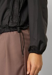 Zwarte lichtgewicht windjack met elastische manchetten en een verstelbare trekkoordonderzoom, gecombineerd met textuur bruin broek. Gladde stofafwerking.