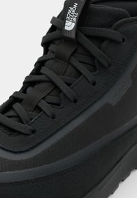 Baskets noires avec une tige synthétique texturée, lacets plats noirs et une semelle en caoutchouc contrastante, avec un logo "DRYVENT" sur le côté.