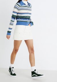 Randig cardigan i blå och gröna nyanser, vit mini kjol och gröna hightop-sneakers med vita accenter. Modellen står.