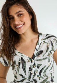 Top de manga corta con base blanca, patrones de hojas verdes y negras, detalles en dorado y escote en forma de V con botones.