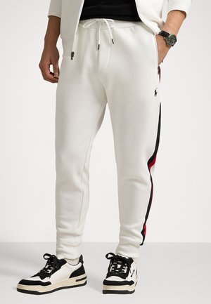 Hvide sweatpants lavet af blødt stof med sidelommer og sorte og røde striber langs benene. Sammen med sorte og hvide sneakers.