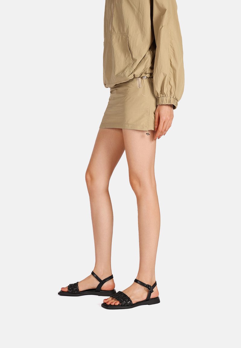 Sandali neri piatti con cinturini intrecciati e fibbia regolabile alla caviglia. Abbinati a una gonna elastica beige e a una maglia oversized coordinata.