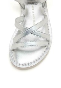 Sandalias metálicas plateadas con correas cruzadas, textura suave, diseño de punta abierta y detalles cosidos en una suela clara.