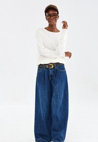 Vrouw in een bruine pet, met grote brillen, een witte lange mouw top en wijde blauwe jeans met een zwarte riem, staande tegen een witte achtergrond.