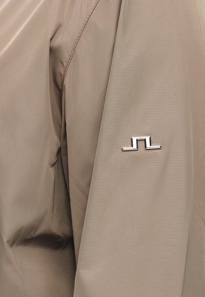 Ύφασμα μπεζ μπουφάν με μικρό ασημένιο γεωμετρικό λογότυπο στο μανίκι κοντά στον αγκώνα.