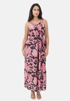 Kvinde står iført en sort maxikjole uden ærmer med et pink blomsterprint og et bindebånd i taljen, kombineret med sandaler med klare stropper.