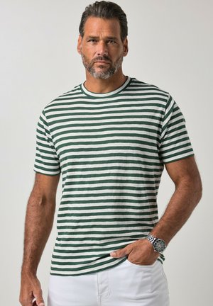 Uomo con capelli corti e barba, che indossa una maglietta a righe verdi e bianche, pantaloni bianchi e un orologio da polso d’argento, con una mano in tasca.