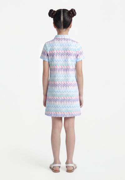 Κορίτσι με δύο κότσους, που φοράει φόρεμα με κοντά μανίκια και μήκος μέχρι το γόνατο, με παστέλ ζιγκ-ζαγκ μοτίβο και λευκά σανδάλια, που στέκεται αντιμετωπίζοντας προς τα πίσω.
