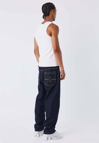 Camiseta sin mangas blanca, vaqueros anchos azul oscuro con costuras de patrón de estrellas en los bolsillos y zapatillas deportivas blancas.