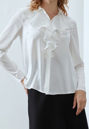 Mujer con blusa blanca de manga larga con detalles fruncidos en la parte delantera y falda negra, de pie con una mano en la cadera.