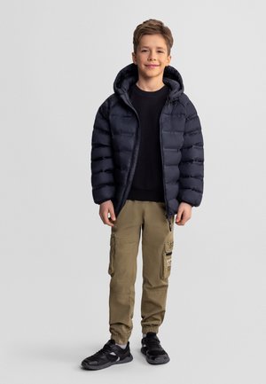 Chłopiec uśmiechający się, ubrany w czarną pikowaną kurtkę, czarną koszulę, khaki spodnie typu cargo i czarne trampki, stojący na tle jednolitego szarego tła.