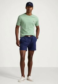 Άνδρας φορώντας κίτρινη μπλούζα, σκούρες μπλε σορτς με ζώνη, λευκά αθλητικά παπούτσια και σκούρο μπλε καπέλο, στέκεται με τα χέρια στις τσέπες.