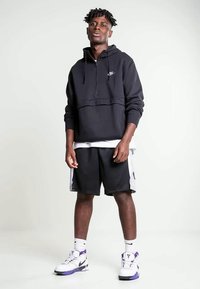 Mustat vetoketjuhupparit, joissa on harmaa yksityiskohta rinnassa, yhdessä mustien shortsejen ja valkoisten lenkkareiden kanssa, joissa on violetteja yksityiskohtia.