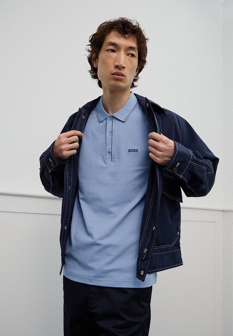Dunkelblaue Jeansjacke über einem hellblauen Polohemd mit Kragen, das ein dezentes "BOSS"-Logo zeigt, vor einem neutralen Hintergrund präsentiert.