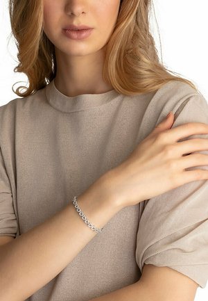 Silberne Kettenarmband an einem Handgelenk, mit ovalen Links und einer glatten Oberfläche. Über einem hellbeigen, kurzärmligen Oberteil getragen.