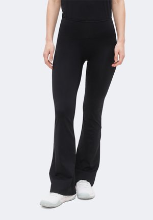 Personne portant un pantalon flare noir ajusté et des baskets blanches, debout avec les mains relâchées le long du corps devant un fond uni.