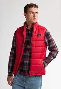 Rote Steppweste mit gestepptem Design, ausgestattet mit einem Stehkragen und seitlichen Taschen, getragen über einem karierten Hemd in Marine, Rot und Weiß.