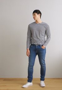 Striped long-sleeve shirt in black and white, paired with blue jeans. Model wears white sneakers, standing against a neutral background.