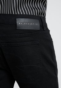 Primer plano de un bolsillo trasero de unos jeans negros que muestra un diseño bordado y una etiqueta de cuero con la inscripción "BALDESSARINI JEANS" en la cintura.